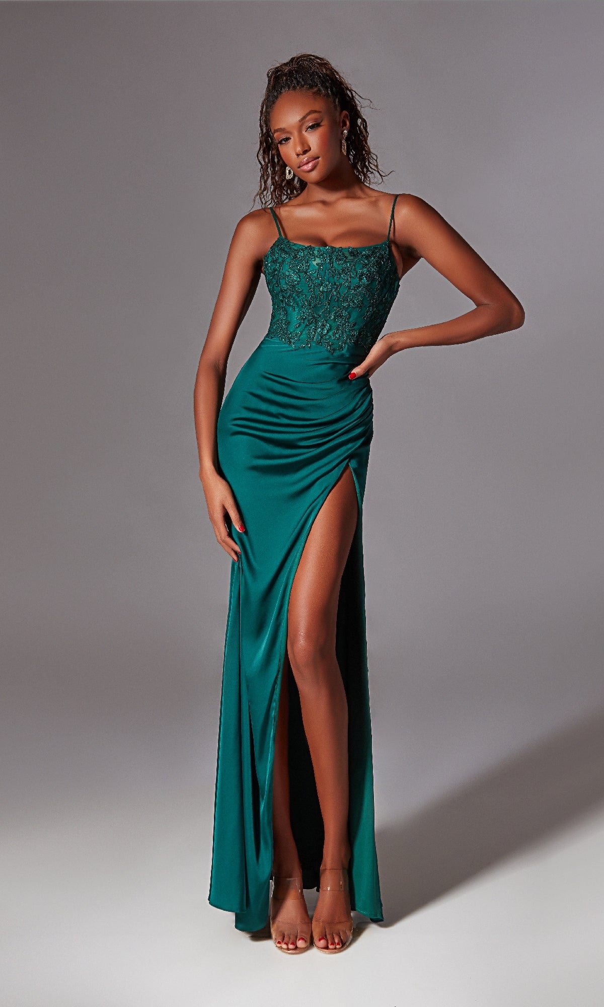 A woman in a Glitter-Bodice Long Prom Dress (Aerie Couture 233), featuring green fabric, sleeveless design, lace bodice, spaghetti straps, high slit, and open back, stands against a plain backdrop.